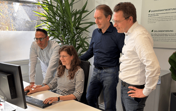 Drei Männer im Büro schauen auf den Bildschirme einer Kollegin