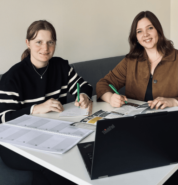 Zwei Mitarbeiterinnen sitzen am Schreibtisch mit Laptop und Kalender