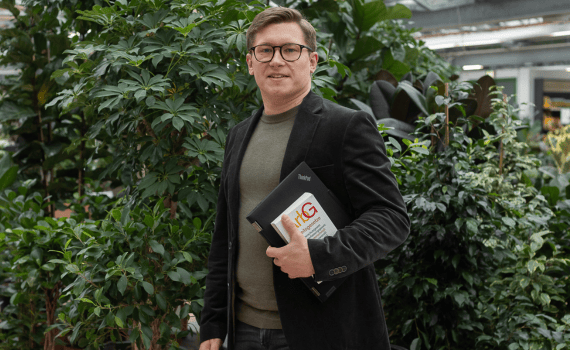 Ein Mann in dunkelgrünem Sakko steht mit Laptop und Gesetzbuch vor einer grünen Pflanze