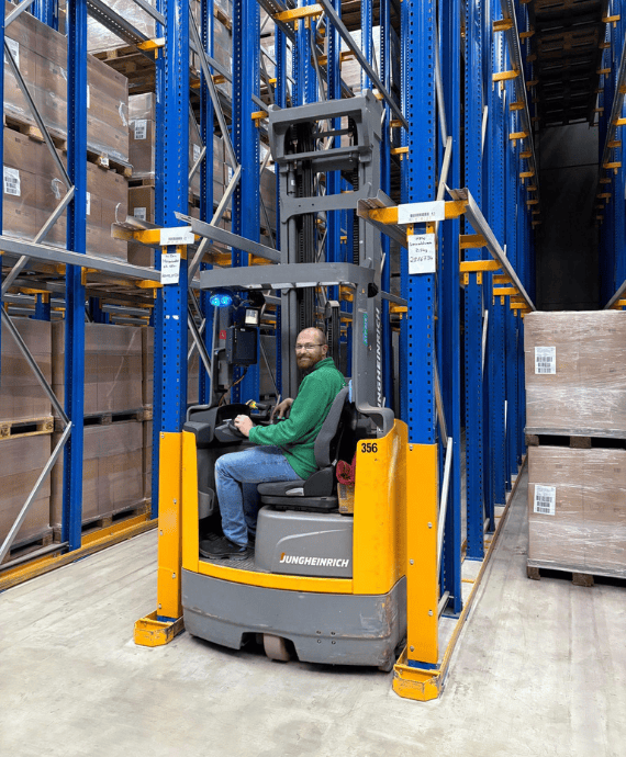 Ein Mann in Arbeitskleidung sitzt auf Stapler und steuert ihn ins Hochregal