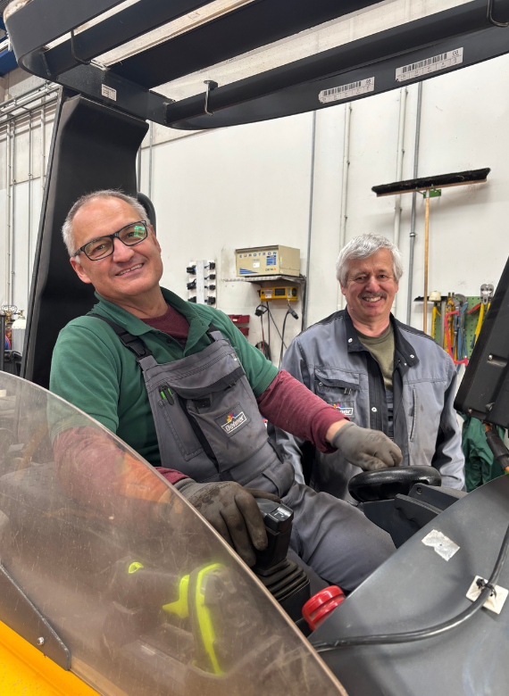Ein Mann in Arbeitskleidung sitzt auf einem Stapler in einer Werkstatt und ein Kollege steht daneben