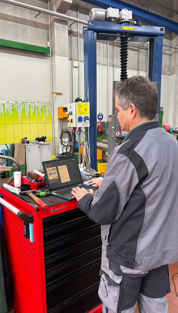 Ein Mann in grauer Arbeitskleidung steht in einer Werkstatt und arbeitet an einem Laptop