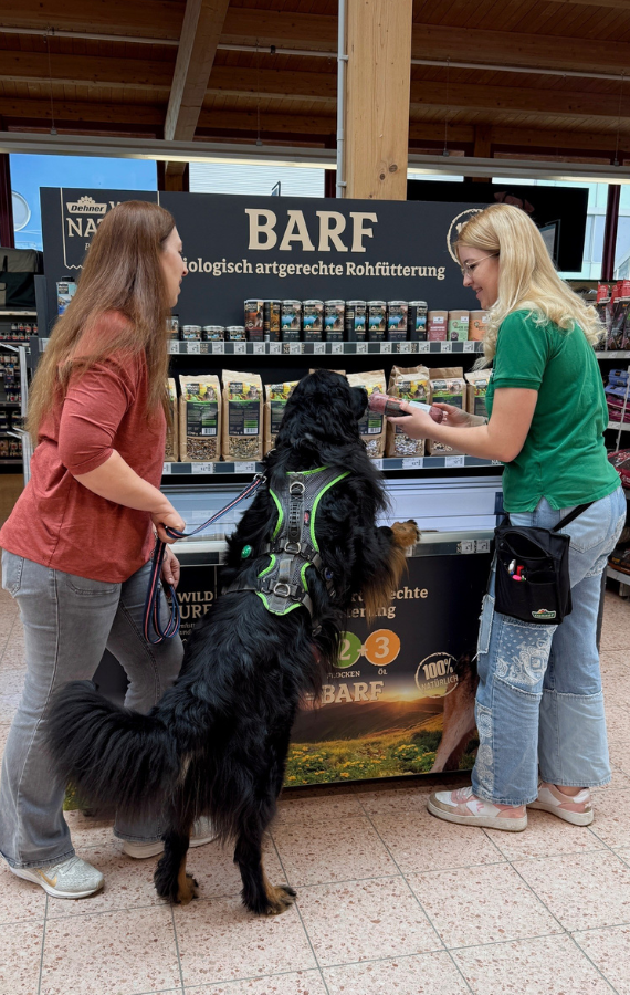 Zwei Frauen und ein Hund stehen vor einer Kühltruhe im Hundefutterbereich und unterhalten sich