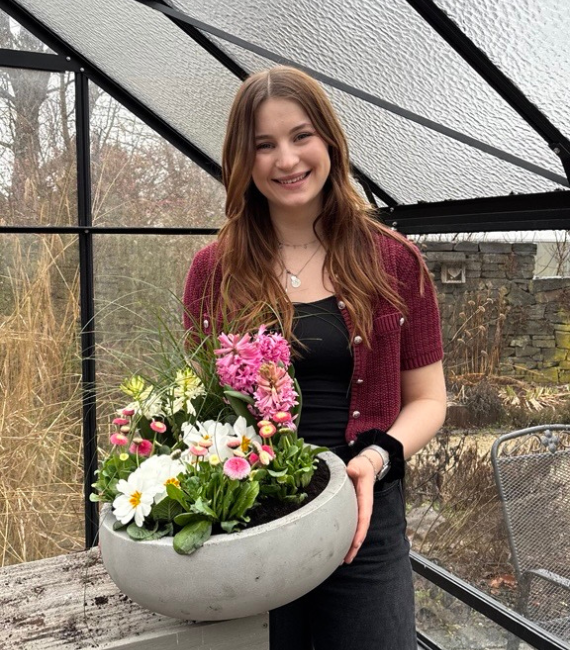 Frau mit roter Strickjacke hält in einem Gewächshaus eine Schale mit eingepflanzten Blumen in der Hand