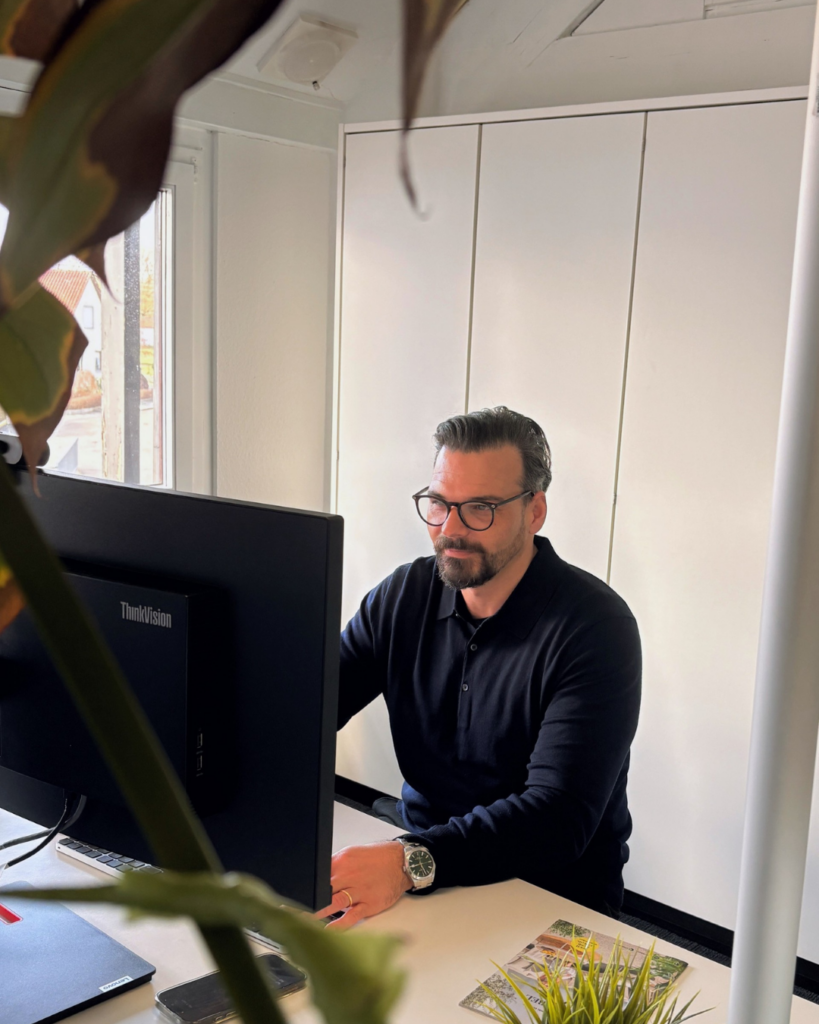 Mann im schwarzen Pullover sitzt am Schreibtisch und liest etwas am PC