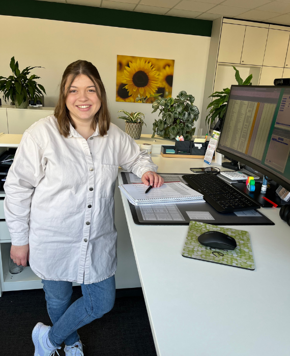 Junge Frau in weißer Bluse steht an einem Schreibtisch und lächelt in die Kamera