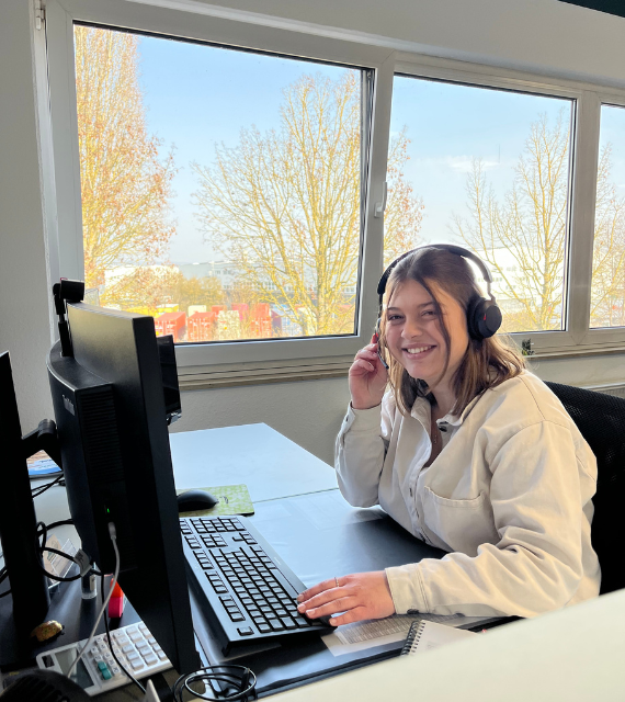 Junge Mitarbeiterin in weißer Bluse sitzt am Schreibtisch und telefoniert mit einem Headset