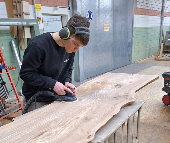 Auszubildender bearbeitet mit einem Schleifer ein Holzbrett