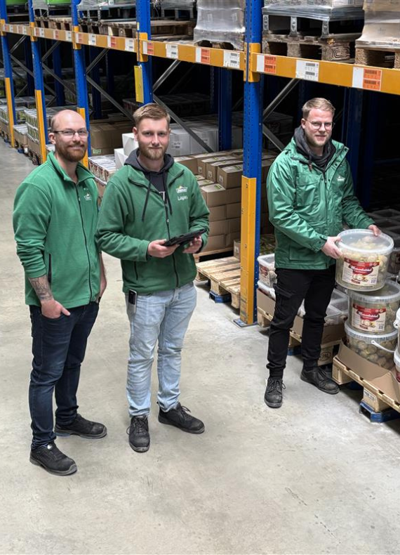 Drei Männer in grünen Jacken stehen zusammen in einem Lager