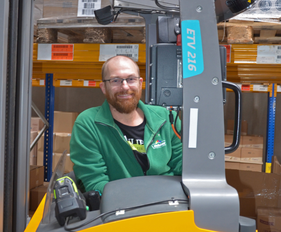Ein Mann in grüner Arbeitskleidung sitzt freudig auf einem Gabelstapler bei der Arbeit