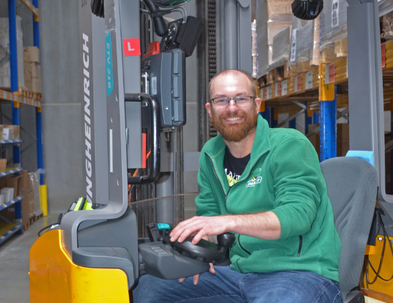 Ein Mann in grüner Jacke sitzt lächelnd auf einem Gabelstabler in einem Lager