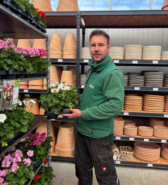 Ein Mann in grüner Jacke steht vor einem Regal mit bunten Geranien