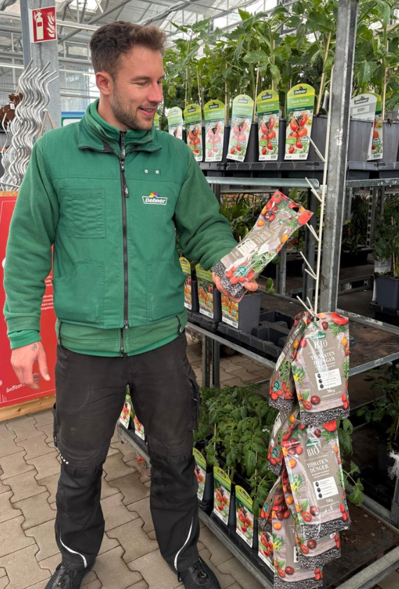 Ein Mann in grüner Jacke steht vor einem Rollwagen mit Tomatenpflanzen und hält einen Dünger in der Hand