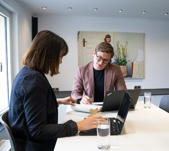 Eine Frau im blauen Blazer und ein Mann im rosa Sakko sitzen an einem Tisch und blättern gemeinsam in einem Gesetzbuch