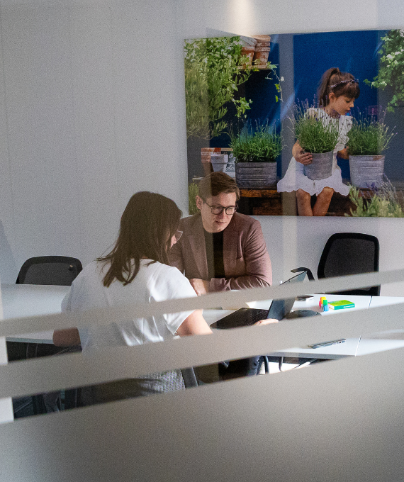 Eine Frau in weißem Shirt und ein Mann in einem rosa Sakko sitzen in einem Meetingraum und erarbeiten gemeinsam etwas an einem Laptop