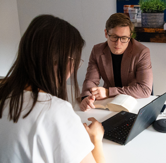 Eine Frau und ein Mann mit einem Gesetzbuch sitzen in einem Besprechungsraum und schauen gemeinsam in einen Laptop