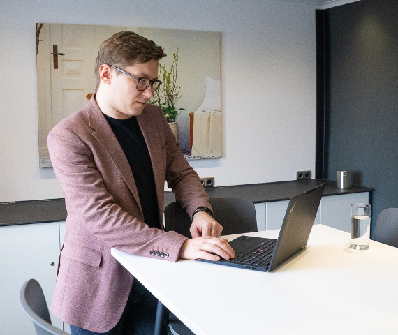 Mann in rosa Sakko steht an einem Tisch und blickt vertieft in seinen Laptop