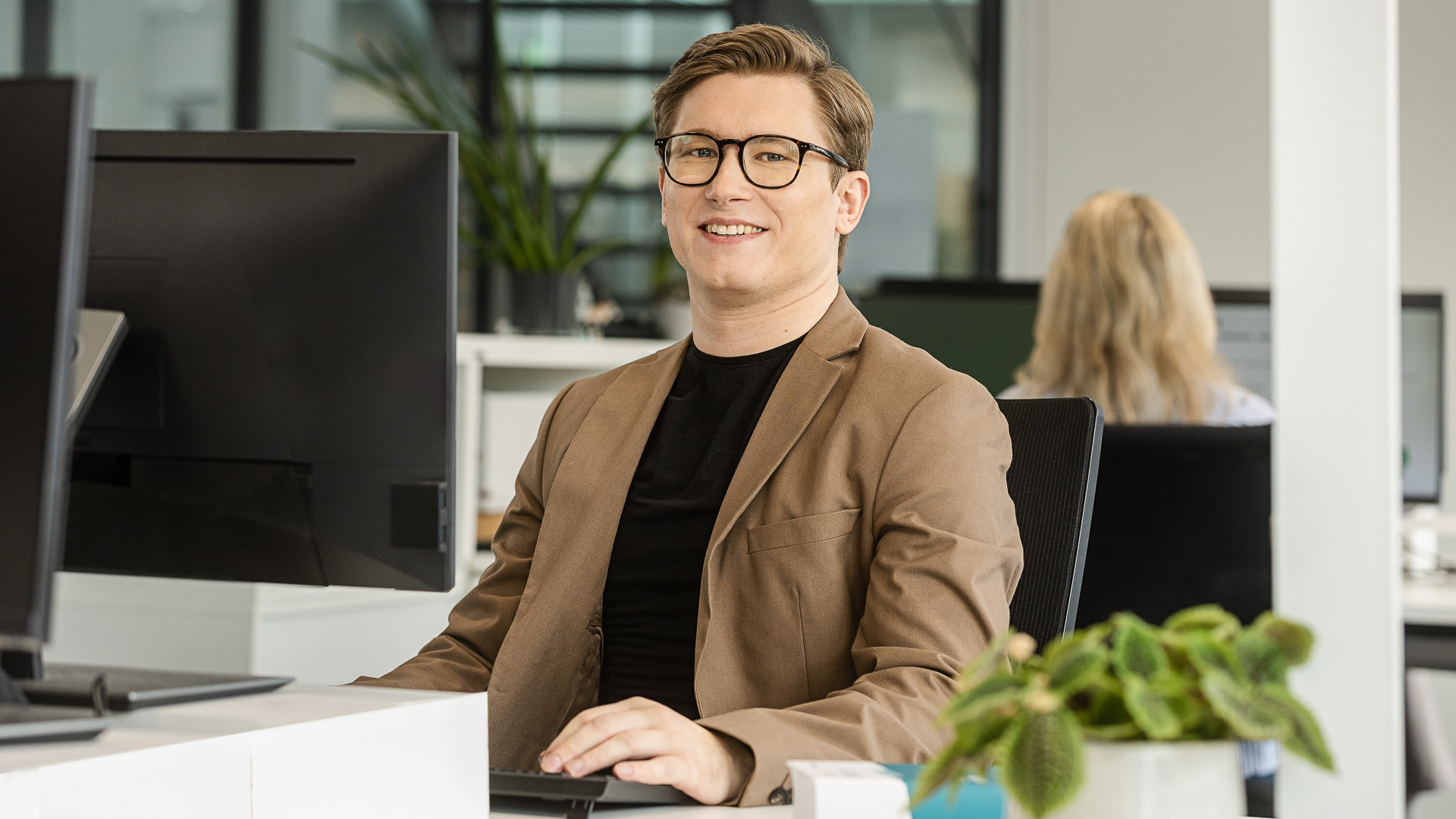 Mann in braunem Sakko sitzt lächelnd im Büro
