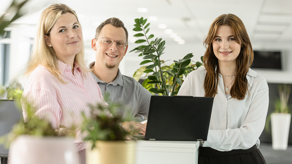 Zwei Frauen und ein Mann stehen in einem Büro lächelnd zusammen und sind von grünen Pflanzen umgeben