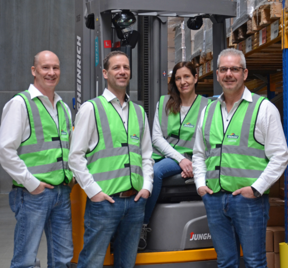 Drei Männer und eine Frau, sitzend auf einem Gabelstapler, tragen Warnwesten und sind in einem Logistiklager