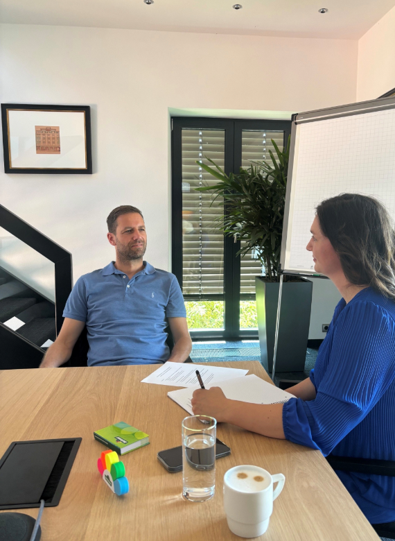Frau in blauem Kleid und Mann in blauem Shirt sitzen an einem Tisch und führen ein Interview