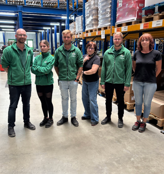 Ein 6-köpfiges Team steht in einem Logistiklager umgeben von Paletten und Ware