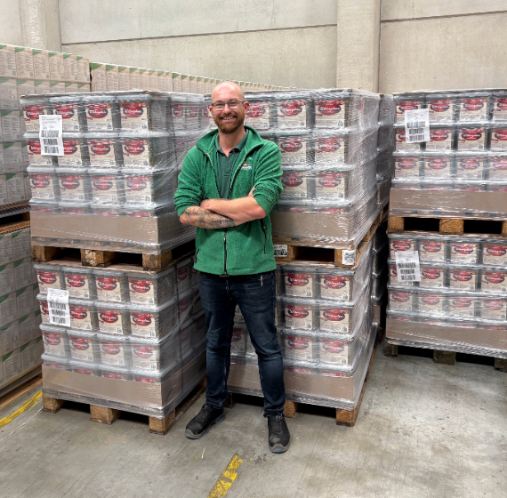 Ein Mann in einer grünen Jacke steht mit verschränkten Armen vor einer Palette mit Wildvogelfutter in einer Logistikhalle