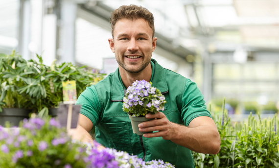 Ein Mann mit grünem T-Shirt hält strahlend eine lilafarbene Blume in einem Garten-Center in der Hand
