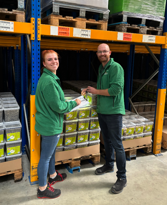 Ein Mann und eine Frau in grüner Arbeitskleidung sind in einem Lager und kontrollieren mit Taschenlampe und Schreibbrett die Qualität der Ware