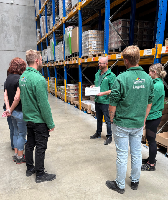 Eine Gruppe aus sechs Mitarbeitenden steht in einem Lager zusammen und hält eine Besprechung
