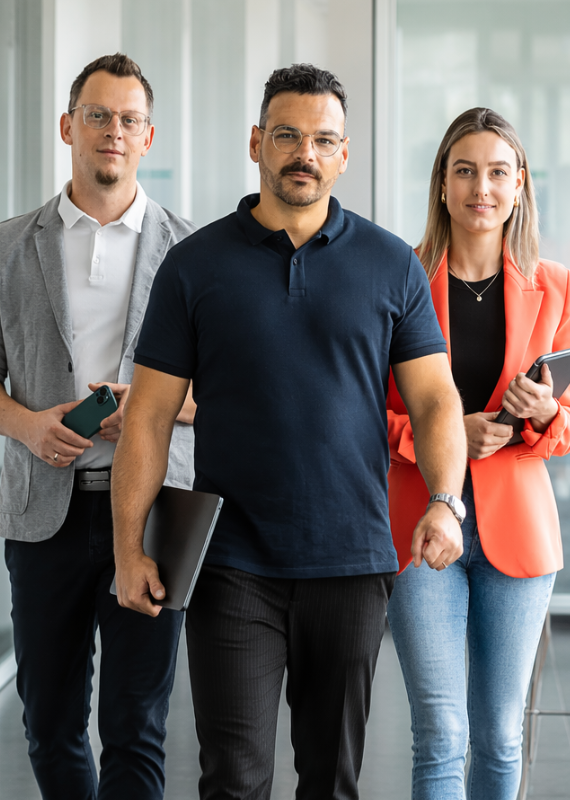 Zwei Männer und eine Frau in Businesskleidung halten Smartphone und Laptop in der Hand und laufen in einer Reihe