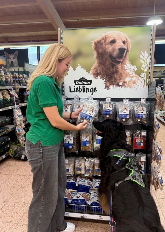 Eine Frau in grünem T-Shirt steht an einem Regal mit Hunde-Snacks und zeigt einem Hund die Packung Rinderohren