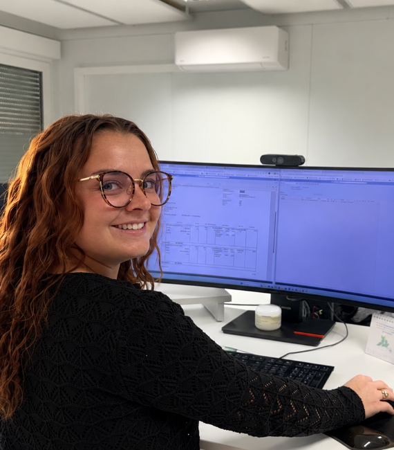 Eine Frau mit langen, lockigen Haaren und Brille arbeitet an einem großen PC-Bildschirm und lächelt